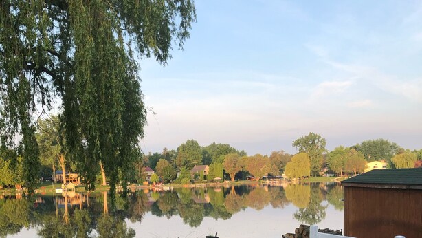 Une terrasse en bois avec deux chaises noires et une table, donnant sur un lac paisible où se reflètent les arbres et les bâtiments.