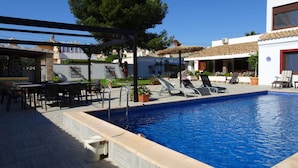 Piscina con sillones de descanso y una mesa, rodeada por un patio con plantas en macetas y una pérgola.