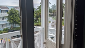 A view from a balcony with a white railing, looking out onto a street with palm trees and a building with a blue roof. - Breezy Studio, footsteps from the beach.  (Negril)