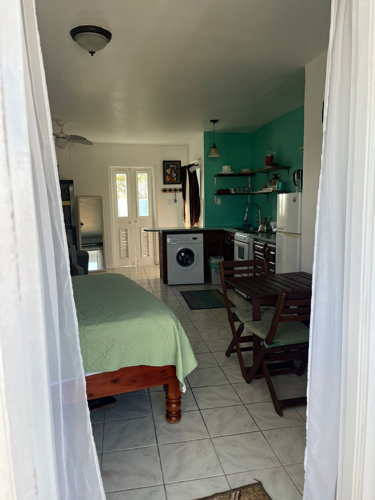 Ein Schlafzimmer mit Bett, Waschmaschine, Esstisch mit Stühlen sowie einer Küchenzeile mit Kühlschrank und Regalen.