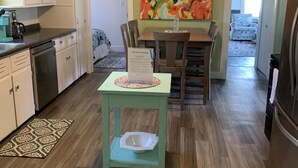 A kitchen with white cabinets, a dining table with chairs, a colorful abstract painting, and a small table with a placemat and a bowl. - Charming House Located In Downtown Grand Haven (Grand Haven)