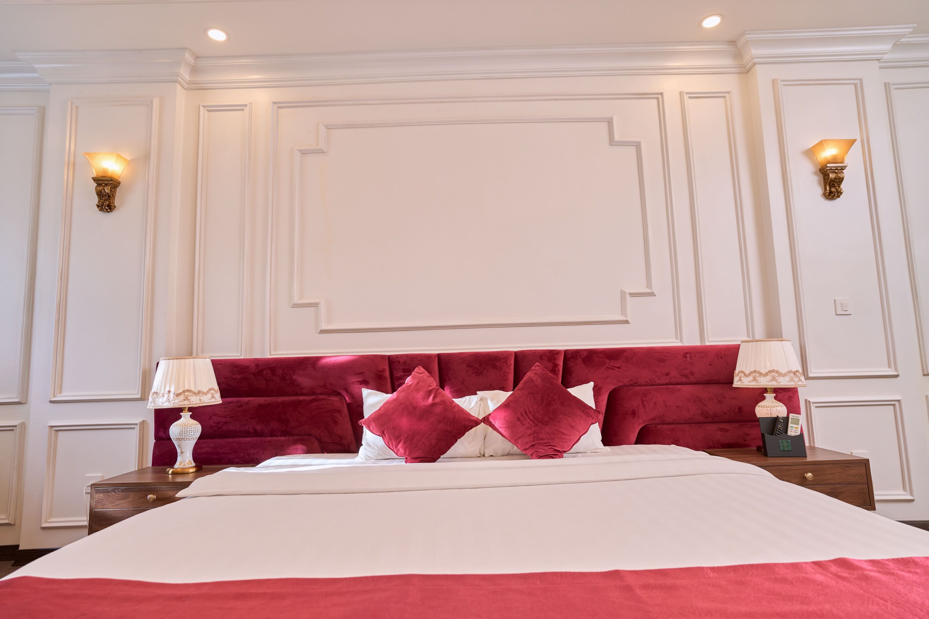 A hotel room with a large bed, red headboard, two bedside tables with lamps, and wall sconces.