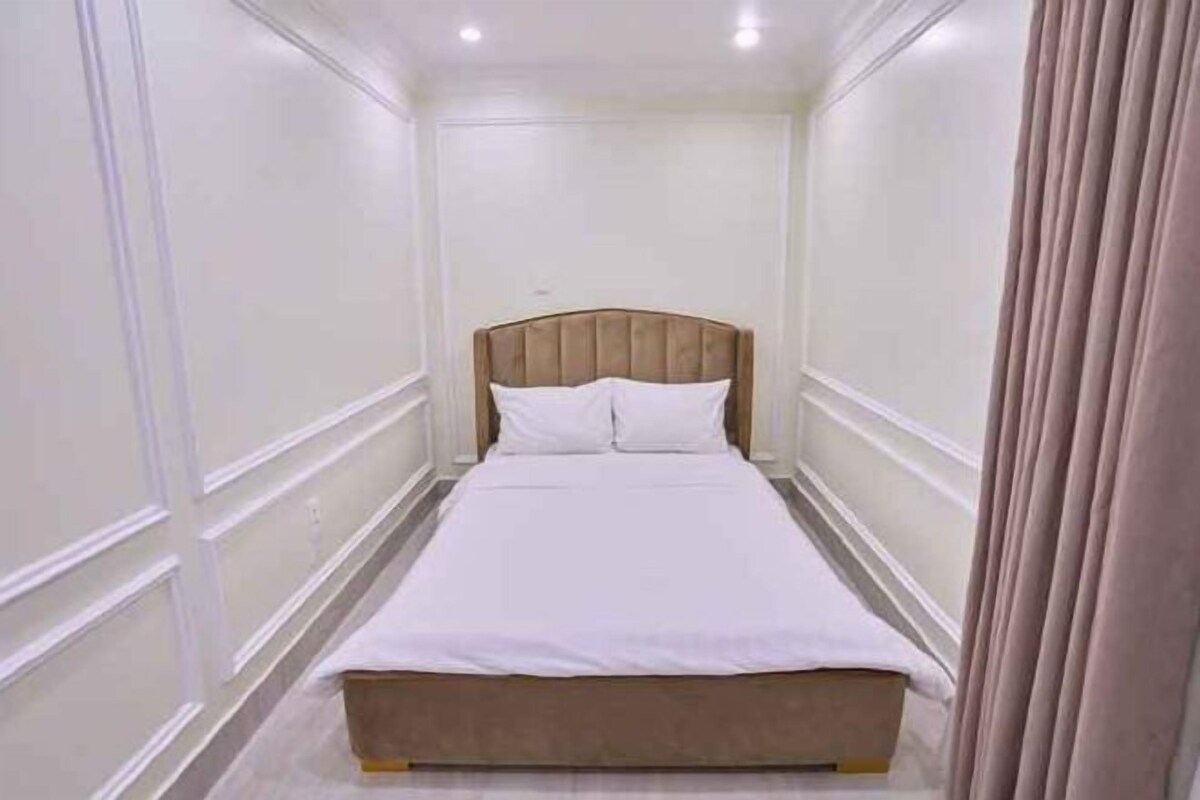 Quarto de hotel estreito com uma cama de solteiro, paredes brancas e cabeceira de madeira.
