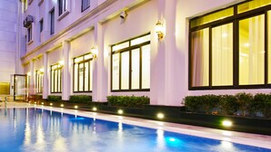 A swimming pool with a reflection of lights, a building facade with large windows, and a row of small bushes. -  Le Haute Hotel Hai Phong (Hai Phong)