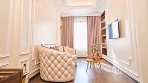 A spacious living room with a large, tufted sofa, a small side table with a vase, and a television mounted on the wall. -  Le Haute Hotel Hai Phong (Hai Phong)