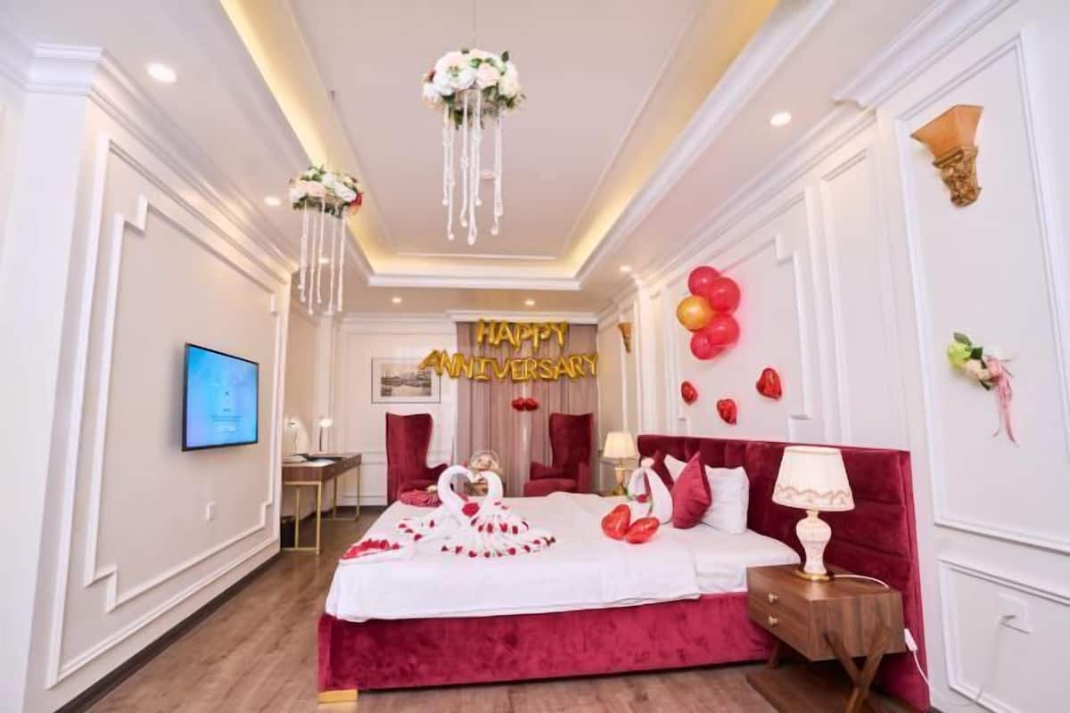 Een hotelkamer met een groot bed, een televisie, een bord met de tekst 'Happy Anniversary' en rode ballonnen.