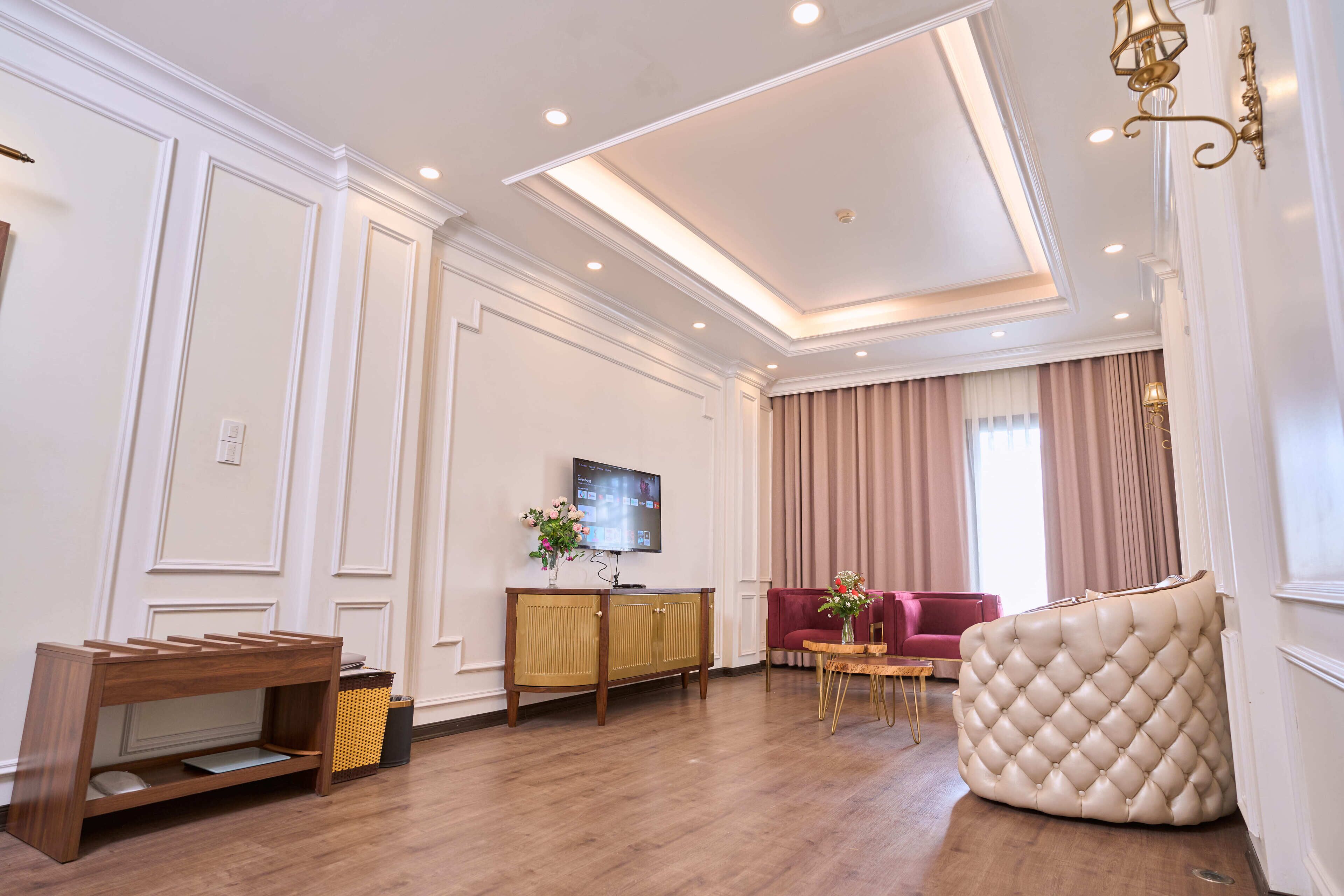 Een ruime woonkamer met houten vloer, een rode bank, een beige poef met knoopjes, een klein tafeltje met een vaas, en een televisie op een houten kast.