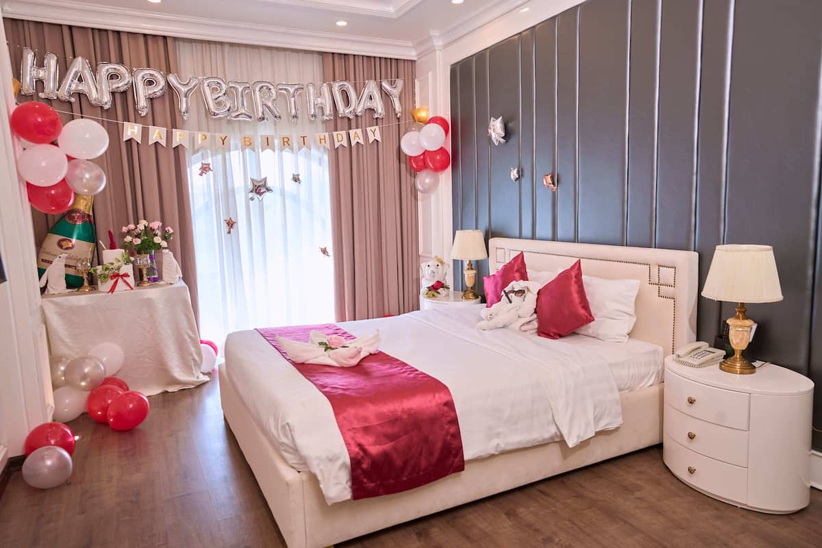 Een ruime slaapkamer met een groot bed, een nachtkastje en een bijzettafeltje. Er is een 'Happy Birthday' banner en ballonnen.