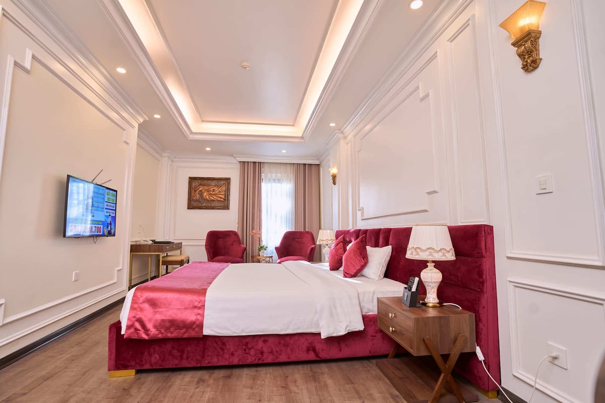Een hotelkamer met een groot bed, twee rode fauteuils, een houten nachtkastje, een televisie en een schilderij aan de muur.