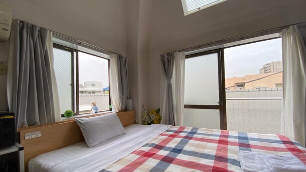 A hotel room with a bed, a window with curtains, a small plant, and a view of buildings outside. - Olympic Park Large Balcony Near Shibuya (Setagaya)