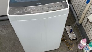 A white Haier washing machine with a control panel, a hose, and cleaning supplies on the floor. - Olympic Park Large Balcony Near Shibuya (Setagaya)