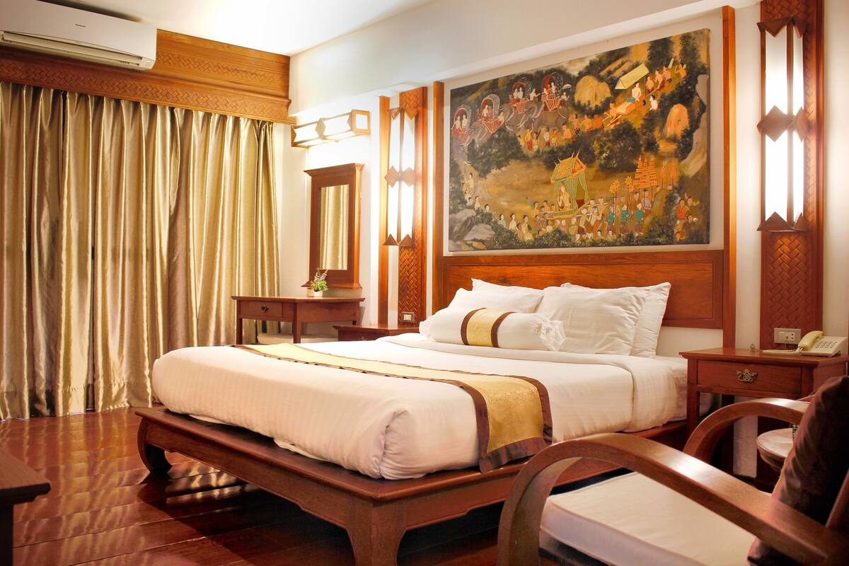 A hotel room with a large bed, wooden headboard, a painting on the wall, a desk with a chair, and a window with curtains.
