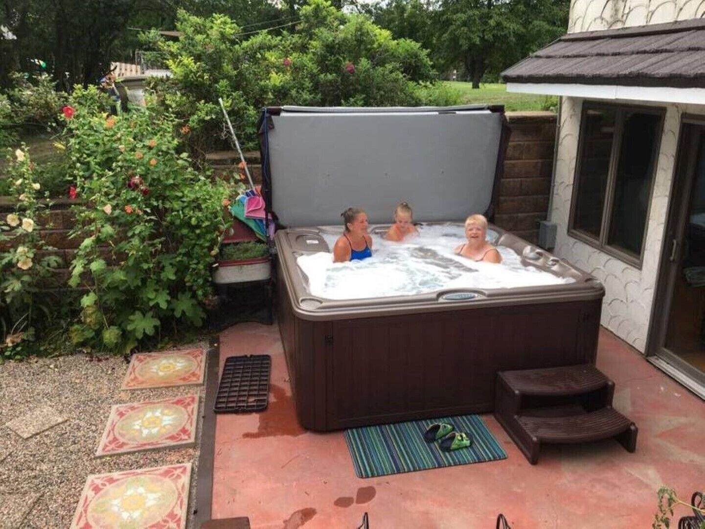 A hot tub with three people in it, surrounded by a garden with plants and a brick wall.