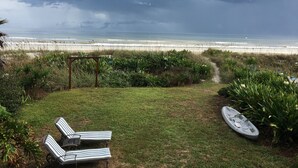 Ein grasbewachsener Bereich mit zwei gestreiften Liegestühlen und einem Kajak, mit Blick auf einen Strand und Wellen.
