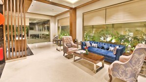 A modern lobby with a blue sofa, patterned armchairs, a wooden reception desk, and large windows with plants. - Hotel Sumaditya (Jaipur)