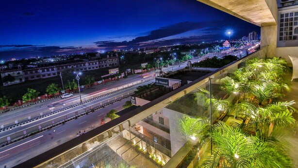 A nighttime view from a balcony overlooking a busy street with a river in the background, streetlights, and a building with a sign that reads "Mumla." - Hotel Sumaditya (Jaipur)