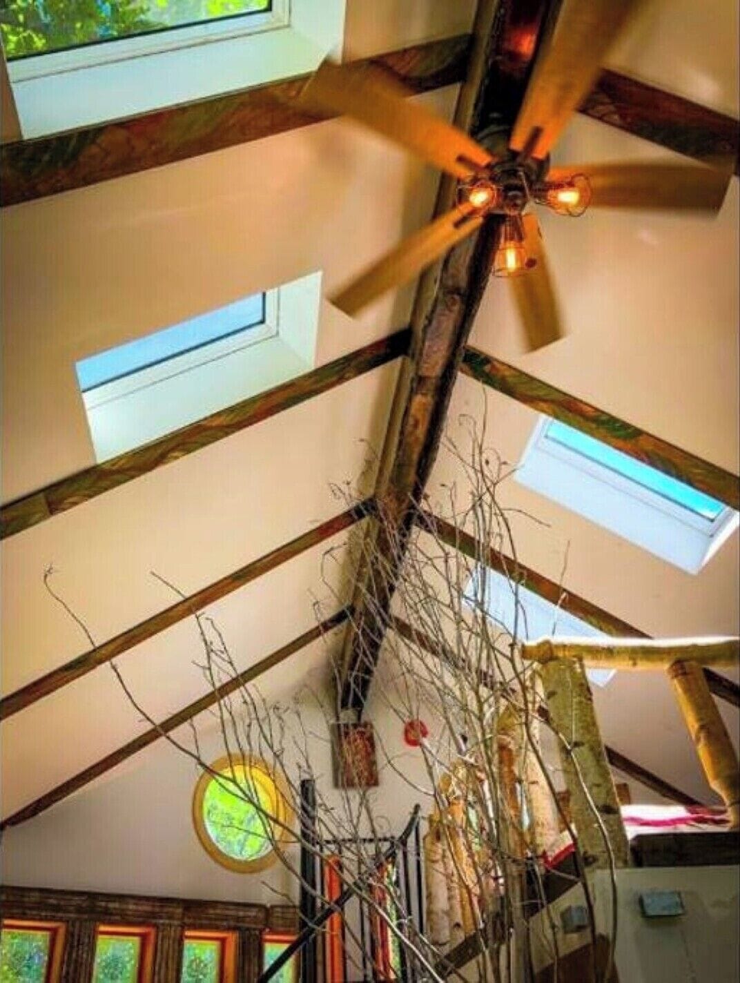 Ein Zimmer mit schrägen Wänden, freiliegenden Holzbalken und einem Deckenventilator. Es gibt Dachfenster und ein rundes Fenster in der Decke.