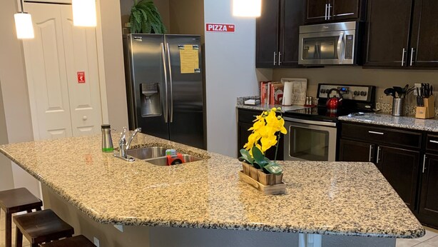A modern kitchen with granite countertops, stainless steel appliances, and wooden bar stools. - 5 Stars House With WiFi, Game Room, Private Heated Spa & Pool In a Gated Area (Kissimmee)