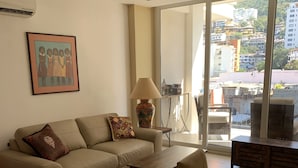 A modern living room with a sofa, coffee table, and dining area. There is a large window with a city view.