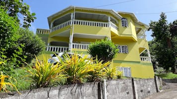 Un edificio amarillo de varios pisos con barandales y balcones blancos, rodeado de follaje y un muro de concreto.