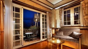 Quarto aconchegante com piso de madeira, mesa de vidro, sofá e vista para um jardim através de grandes portas de vidro.