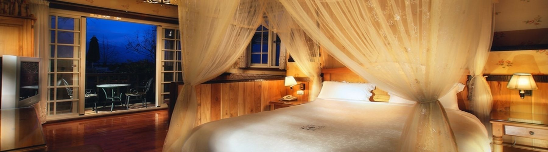 A bedroom with a large bed, a window with curtains, a bedside table with a lamp, and a view of the outside.