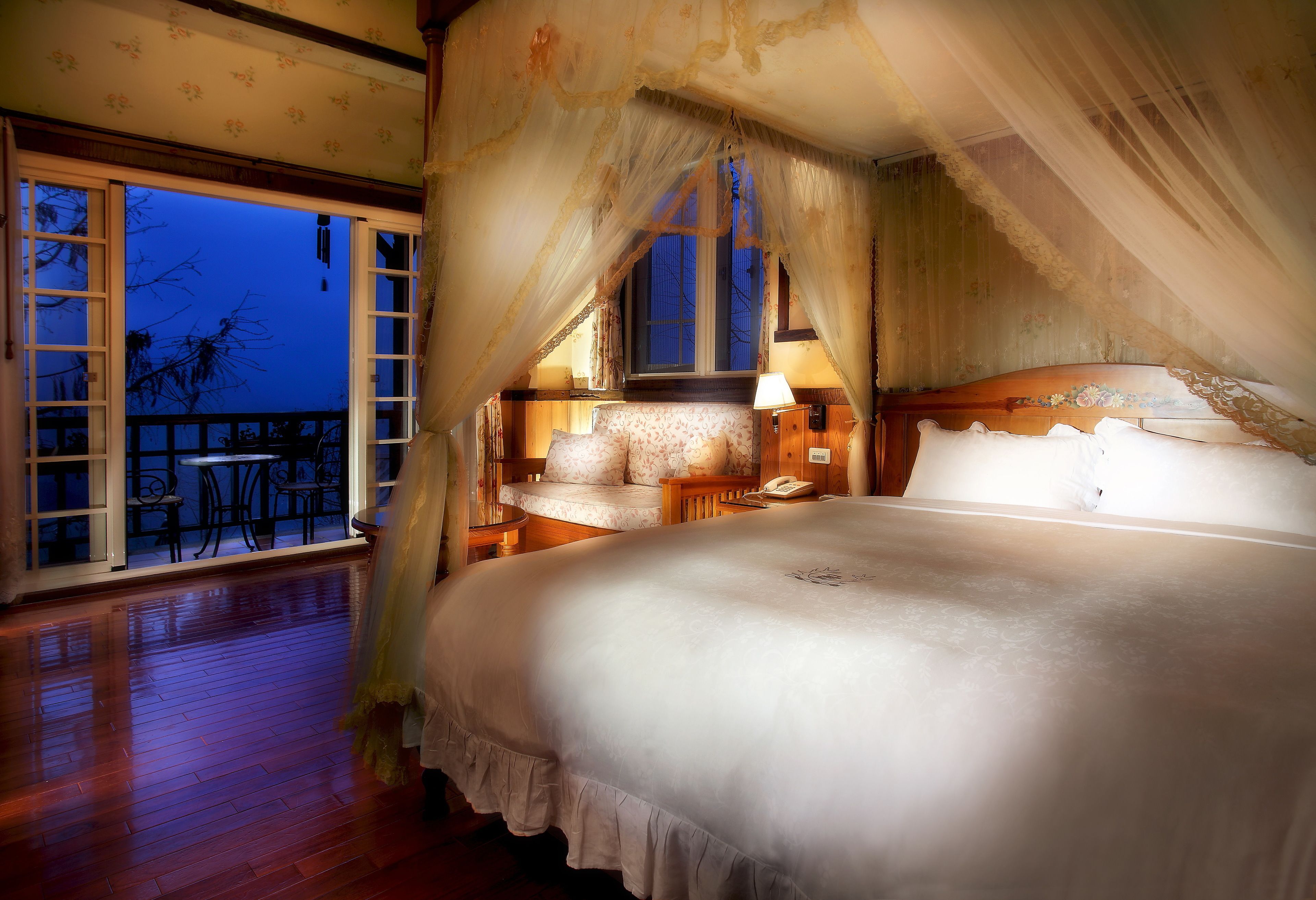 Una cama grande con dosel, piso de madera, balcón con vistas y mesita de noche con lámpara.