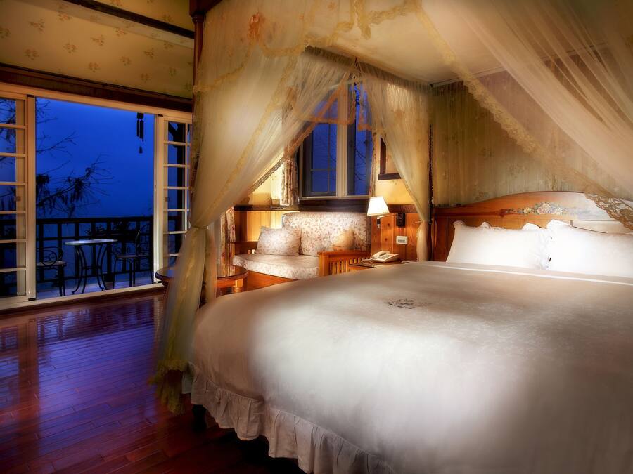 Una cama grande con dosel, piso de madera, balcón con vistas y mesita de noche con lámpara.