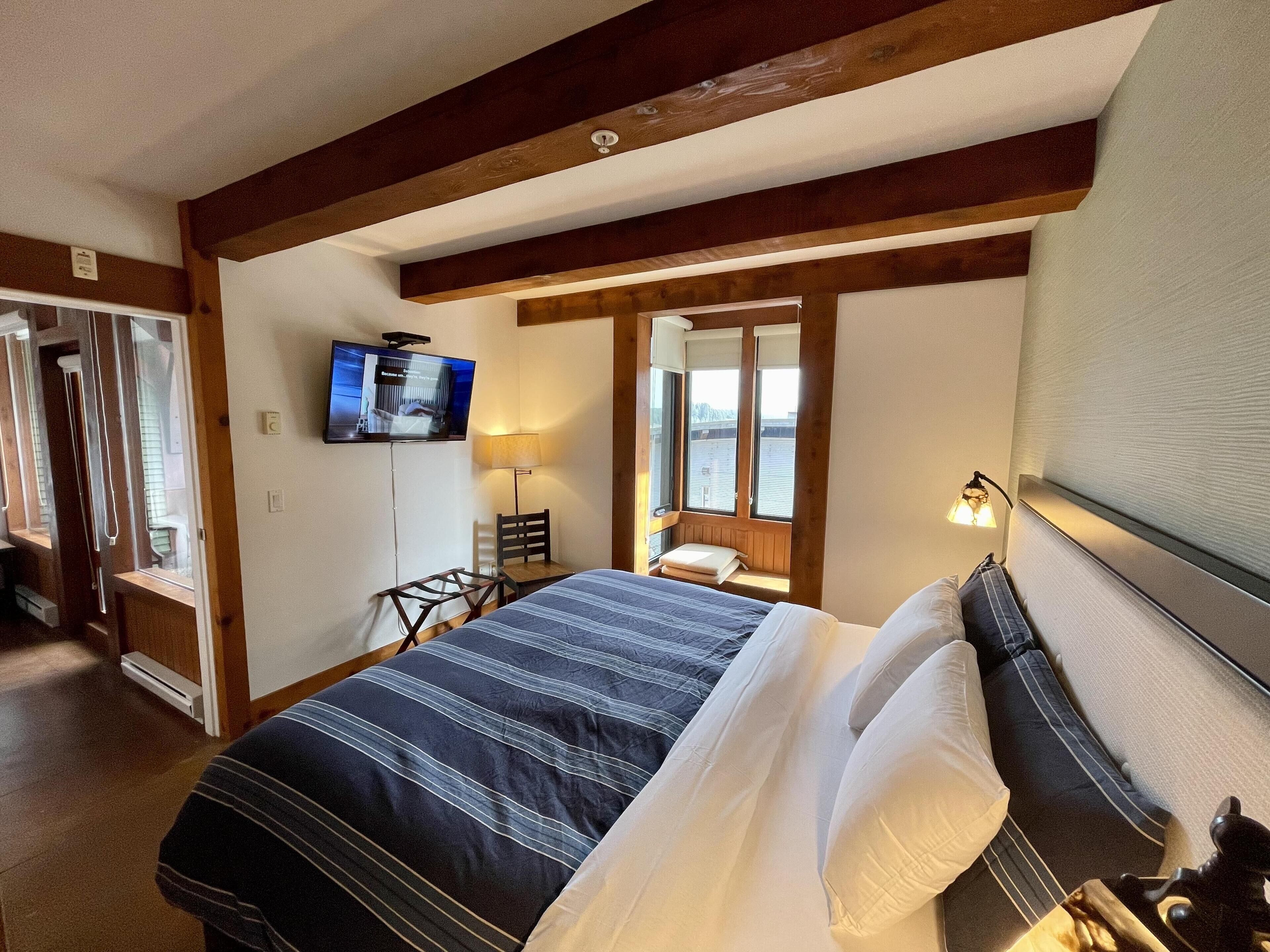 Quarto de hotel com uma cama grande, televisão fixada na parede, um assento junto à janela com abajur e teto de madeira.