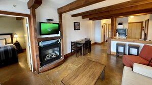 A living room with a fireplace, a television mounted above it, a dining area with a table and chairs, and a kitchen area with a counter and stools.