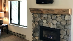 A living room with a stone fireplace, a mounted TV, and a window with blinds.