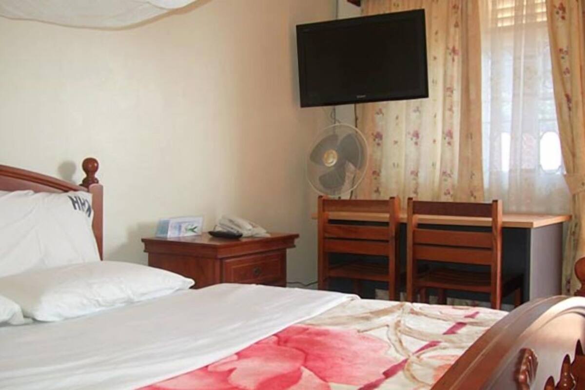 Habitación de hotel con cama, escritorio con silla, televisor montado en la pared y ventilador sobre el escritorio.