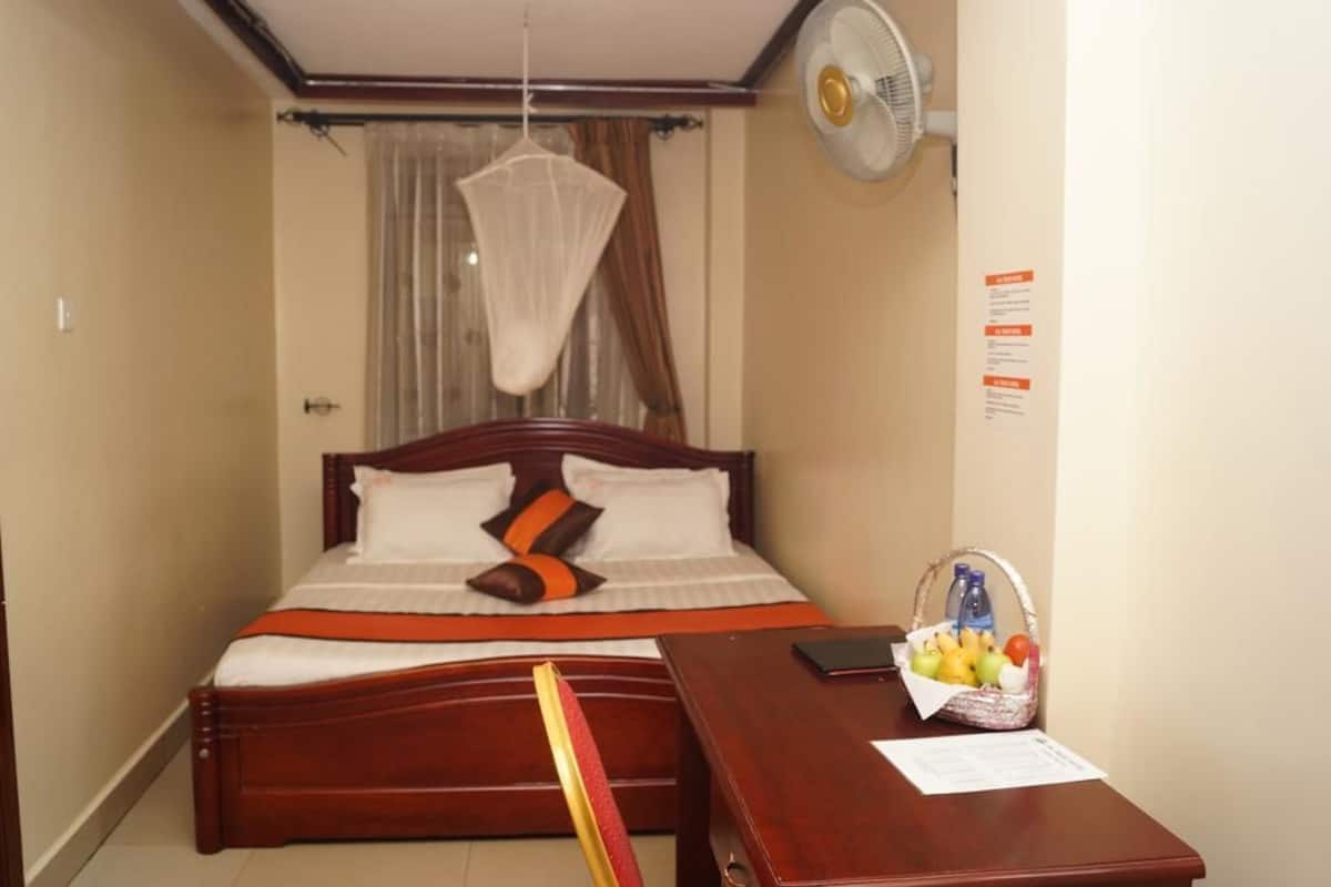 Habitación de hotel con una cama de madera, una mesita de noche con una cesta de frutas, un ventilador y una ventana con cortinas.