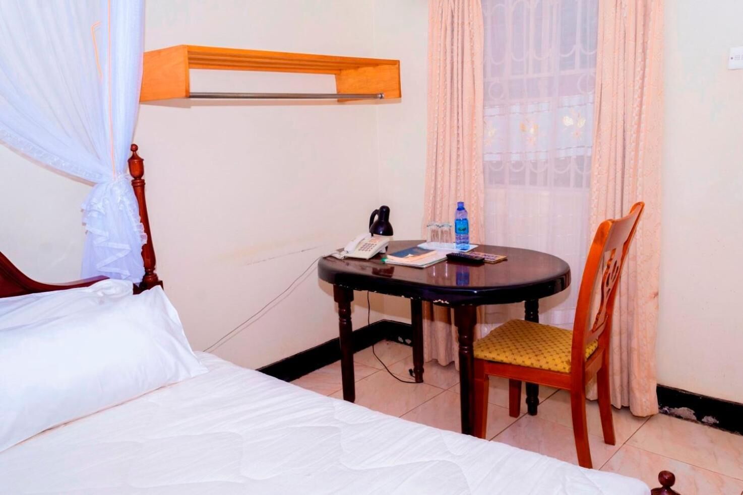 Habitació d'hotel amb un llit, una taula petita amb un telèfon i una ampolla, una cadira i una finestra amb cortines.