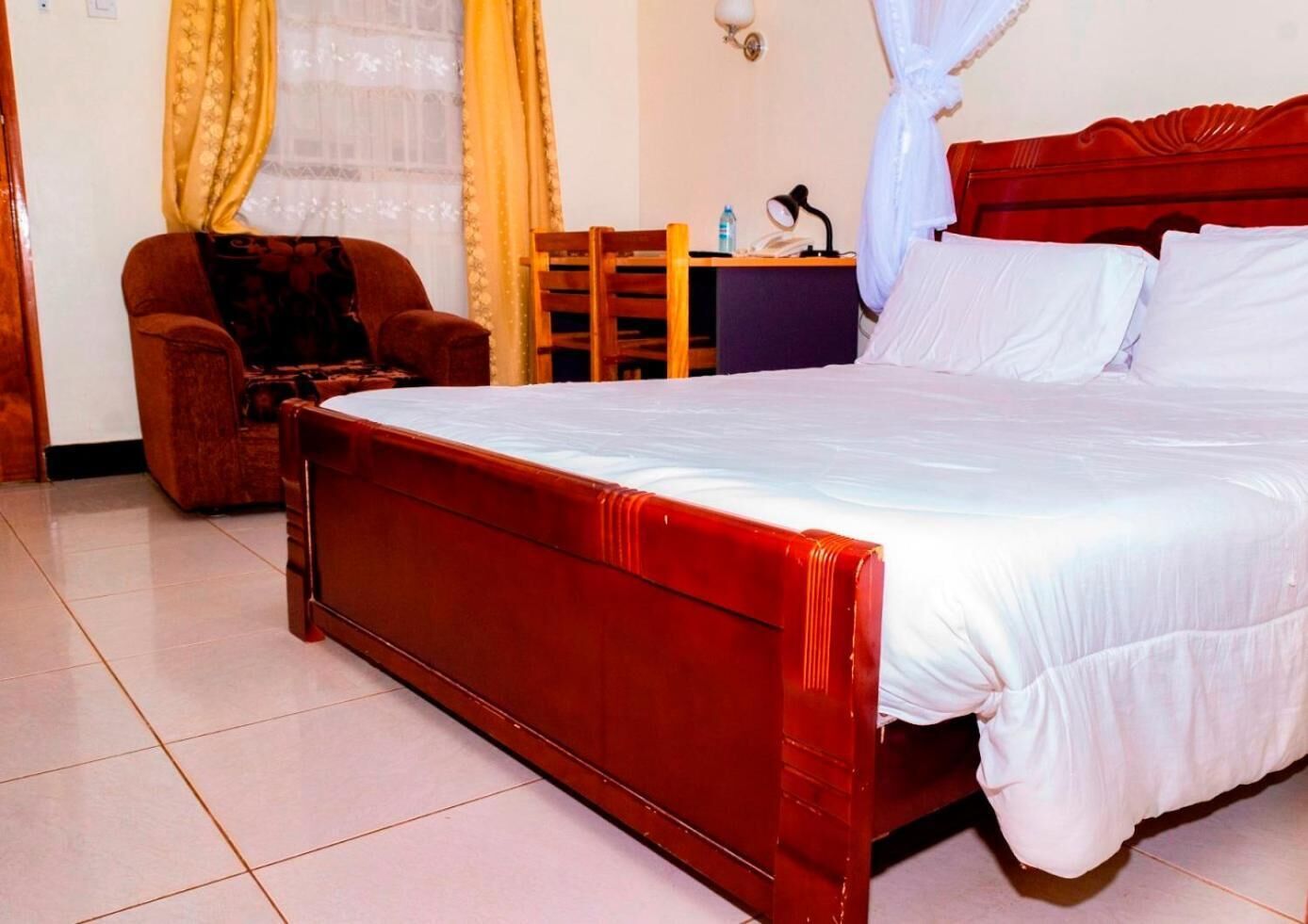 A neatly made bed with a wooden headboard, a chair, a small table with a lamp, and a window with curtains.