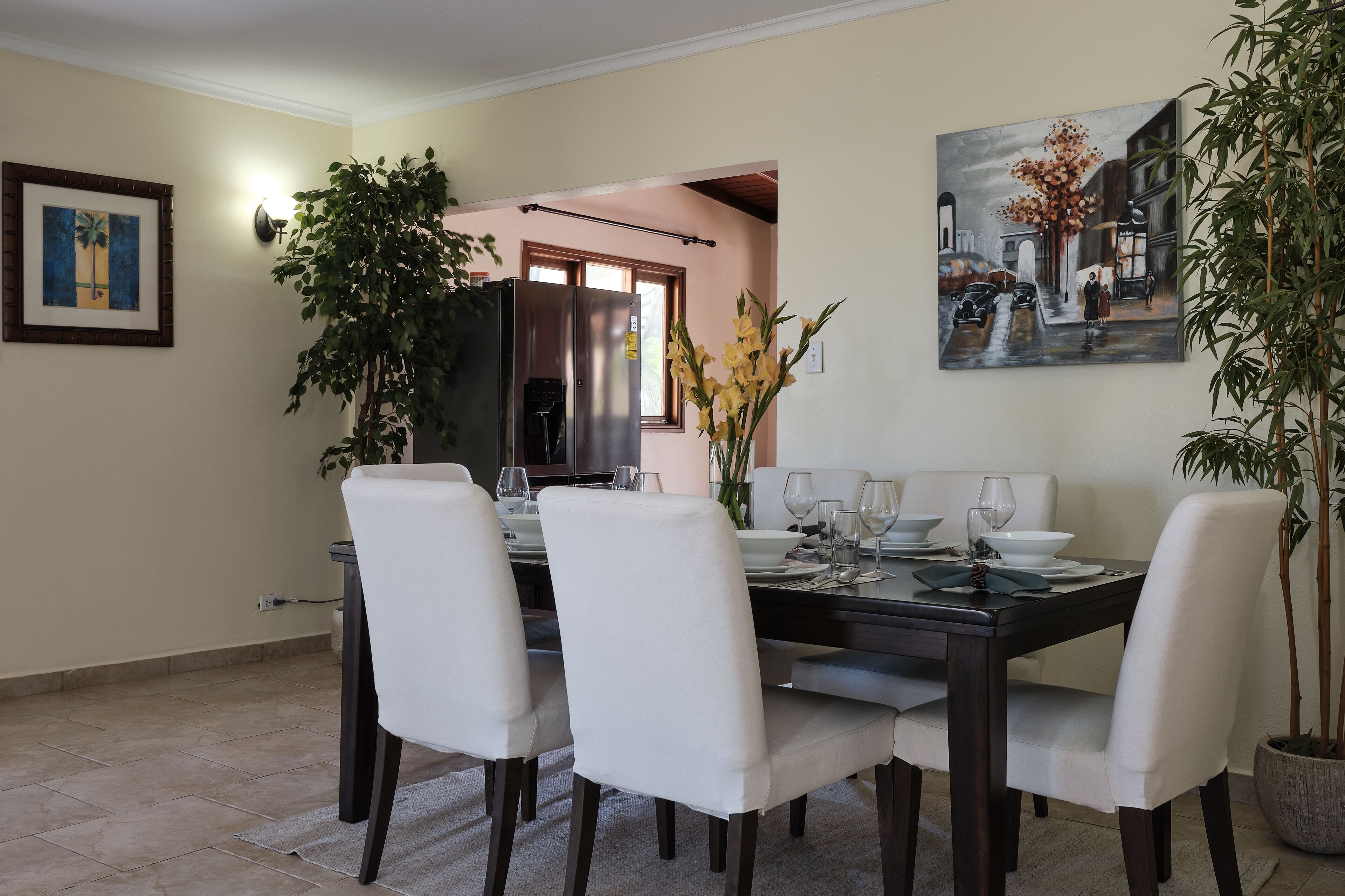 Un coin repas avec une table en bois foncé, des chaises blanches et un tableau représentant une scène de rue.