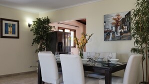 A dining area with a dark wooden table, white chairs, and a painting of a street scene.