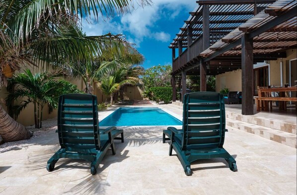 A pool area with two lounge chairs, a pergola, and palm trees.