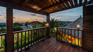 A wooden balcony with a view of a sunset over a landscape with houses and a thatched-roof structure.