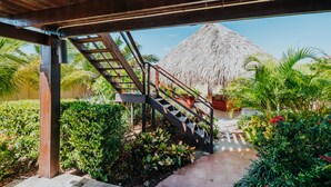 A covered outdoor area with a staircase leading to a thatched-roof structure, surrounded by lush greenery and a tiled pathway.