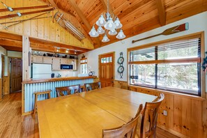 Un interior de madera con una mesa de comedor central, sillas de madera, una zona de cocina con microondas y un ventanal con persianas.