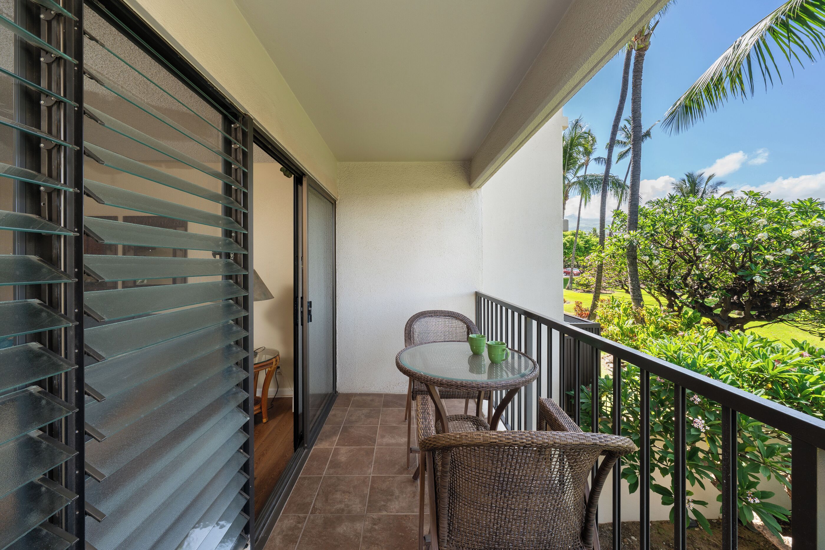 A balcony with a glass door, wicker chairs, a small table, and a view of greenery and palm trees.