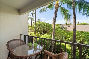 Un balcon doté de meubles en osier et d’une table en verre, offrant une vue sur un jardin luxuriant, avec des palmiers et un bâtiment en arrière-plan.