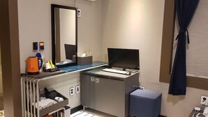 A hotel room with a desk, a flat-screen TV, a mirror, a small seating area, and a window with curtains. - Blue Hotel (Geoje)