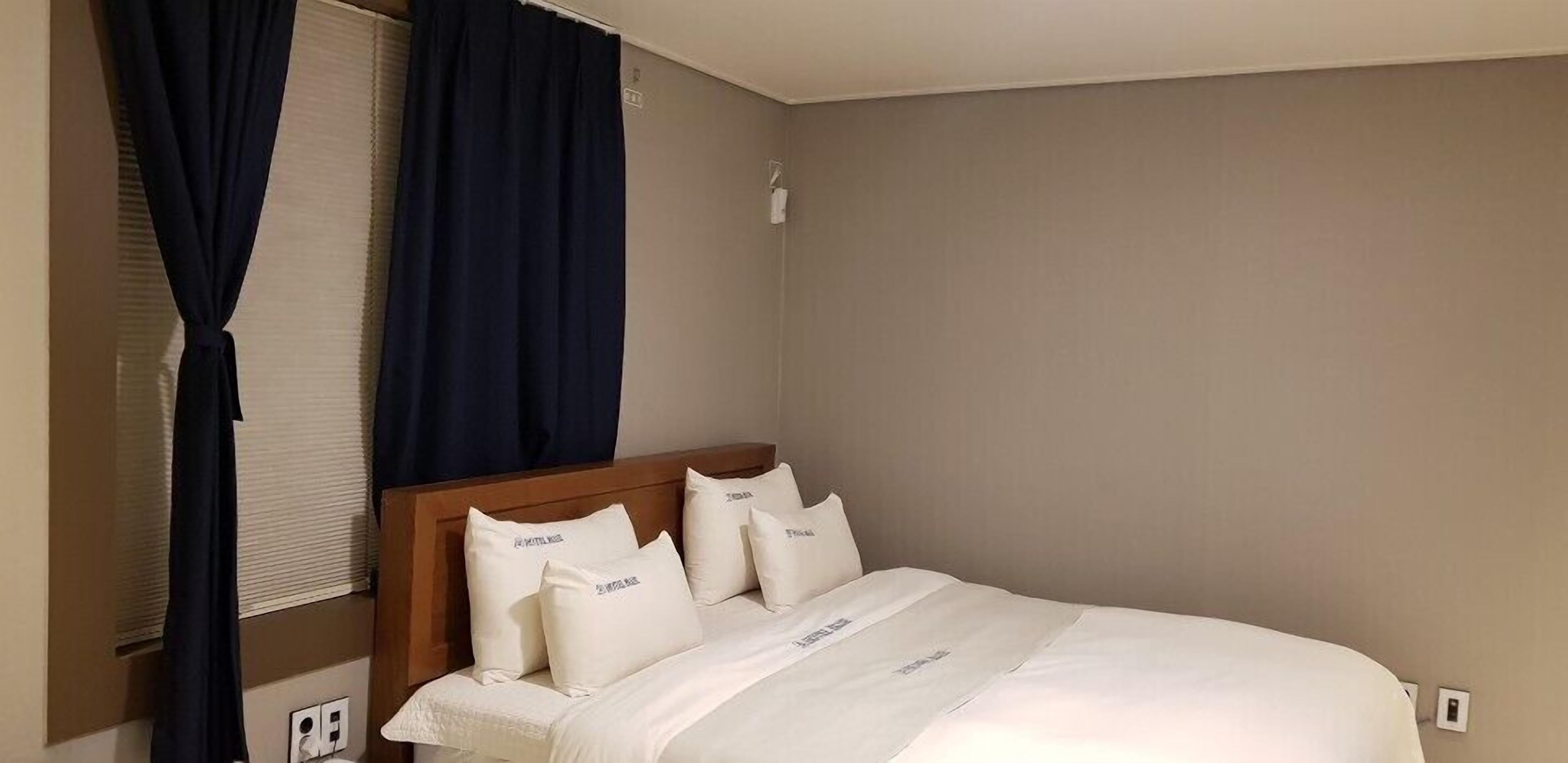 A neatly made bed with white bedding and pillows, a wooden headboard, and a window with dark curtains.