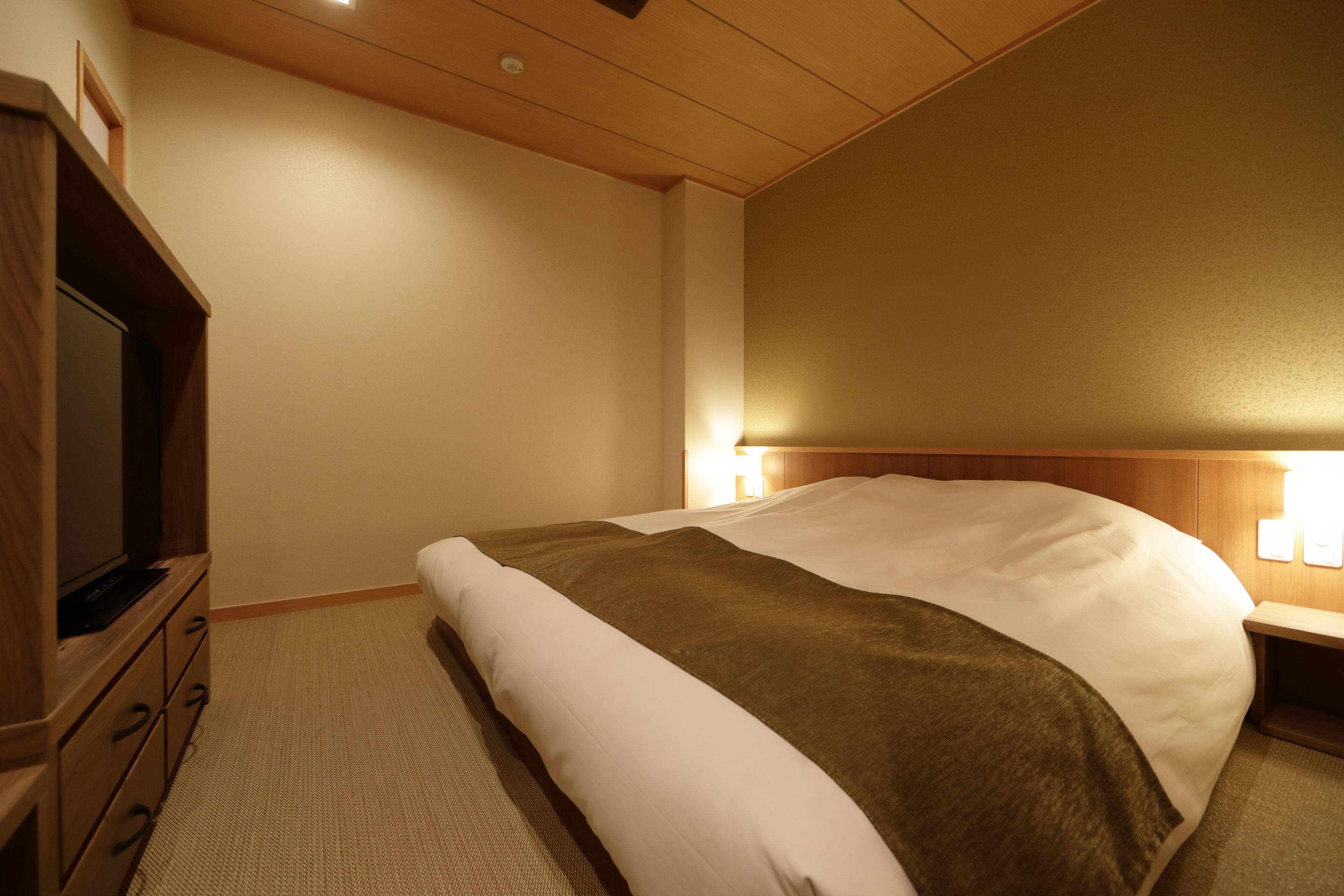 A hotel room with a bed, a TV, a bedside table, and a lamp.