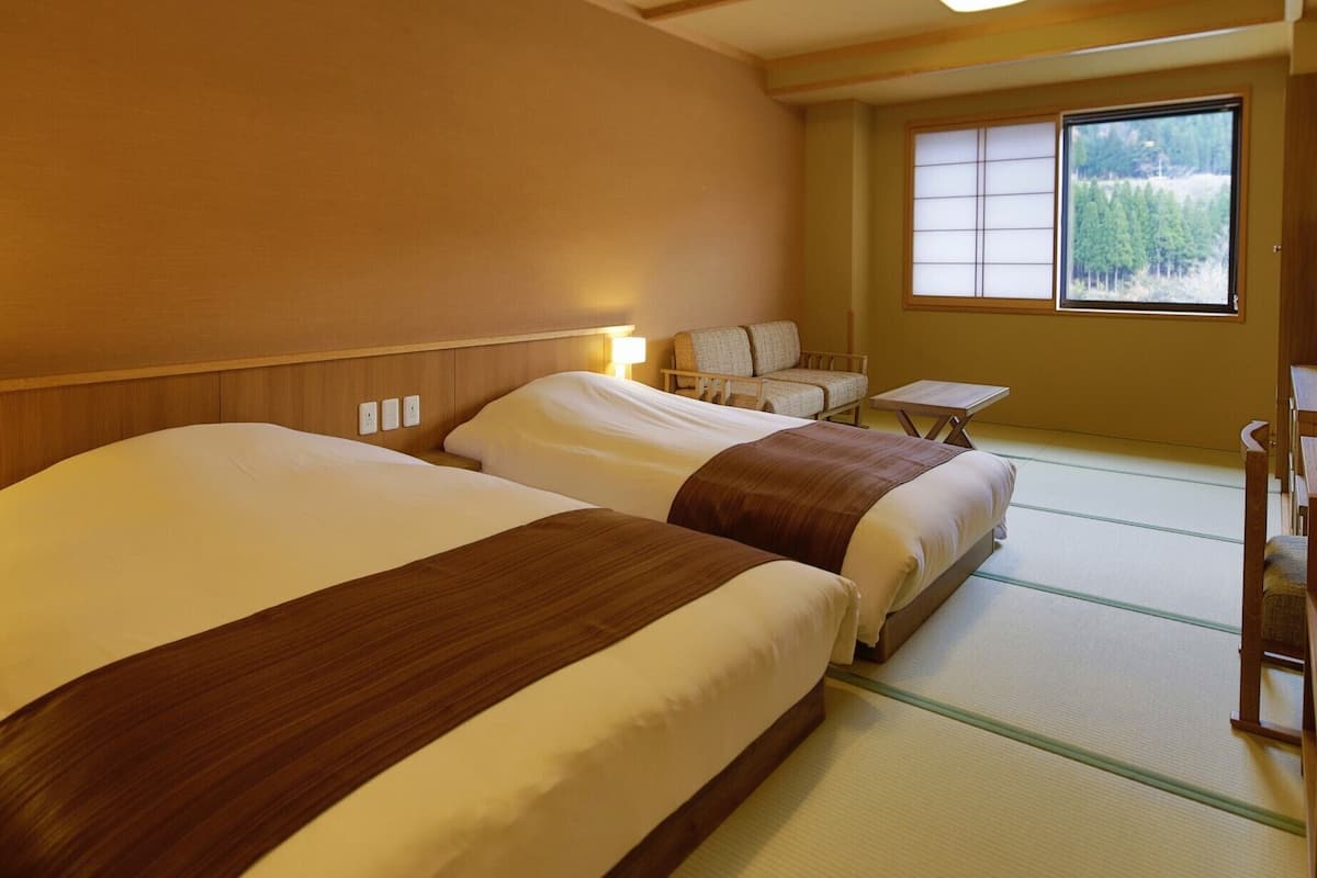A hotel room with two beds, a small table, a chair, and a window with a view of trees.