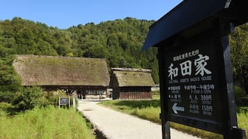 A traditional Japanese house with a thatched roof, surrounded by lush greenery and a clear blue sky.