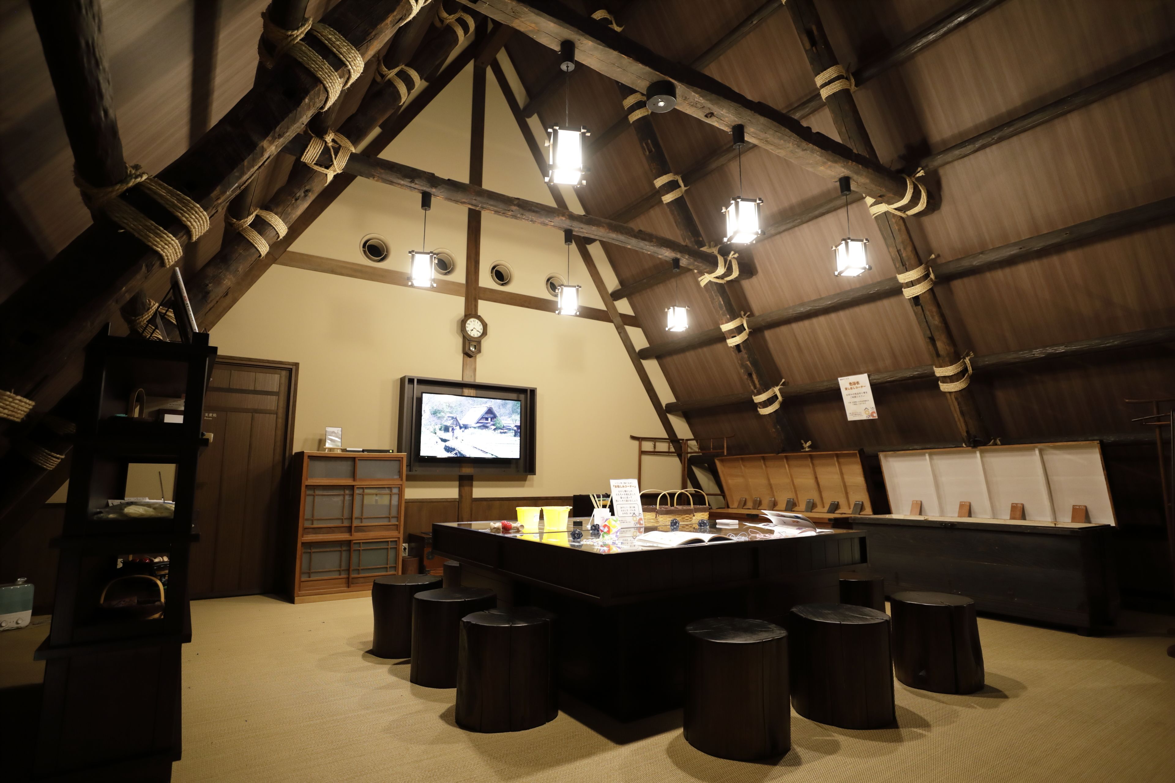 A rustic interior with a wooden ceiling, a central table with stools, a television, and a wooden cabinet.