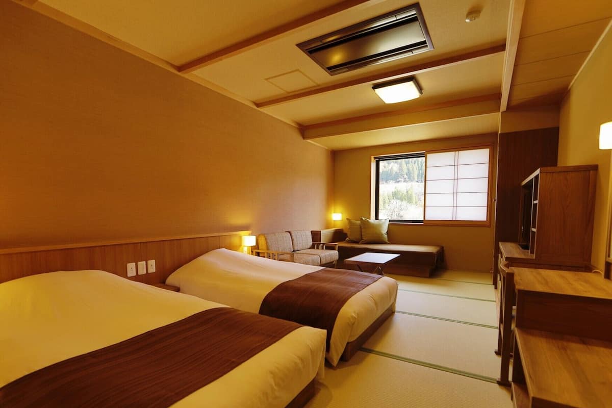 A hotel room with two beds, a sofa, a TV, and a window with a view of trees.